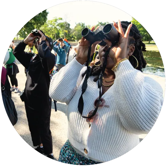 A group of about eight people, including some medium- and light-skinned people, standing on a path on the National Mall and looking up through binoculars. One woman is holding a camera. Some green leafy trees are in the background.