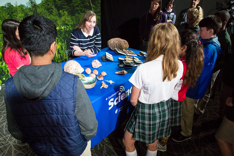 Boys and girls look at snail specimens on a table where zoologist Ellen Strong is sitting. She is speaking to the students.