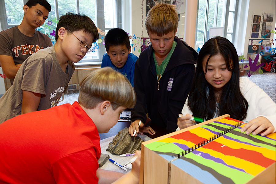 Six teenage students stand around a table with a colored box and rocks on it. They are doing a school program about geology.