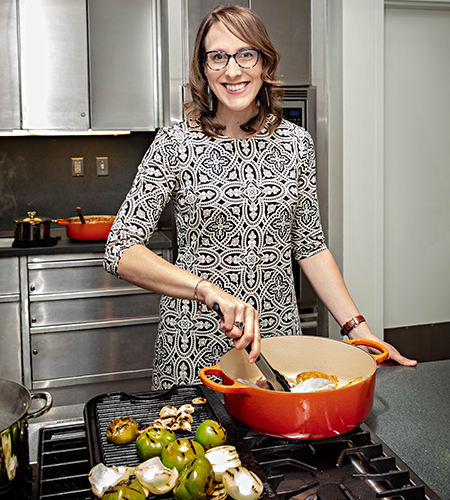 Ashley Rose Young stands over a cooktop, reaching into a pot with a utensil. Next to the pot is an oven tray with vegetables.