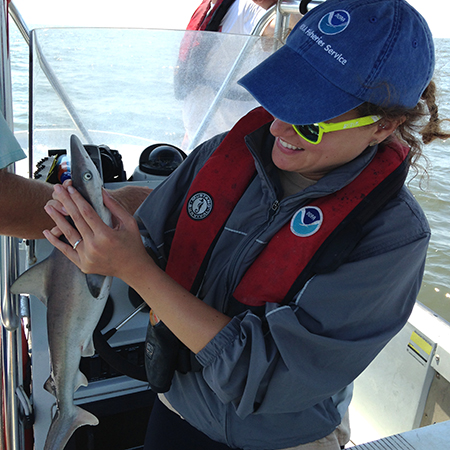 A woman wearing a baseball cap, windbreaker, and life vest, standing on a boat and holding what looks like a baby shark.
