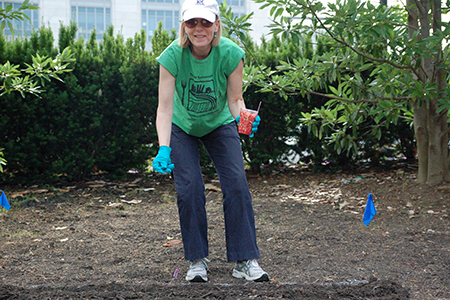 Paula Johnson standing in a garden with a plastic cup full of seeds, getting ready to plant them in a furrow in the ground.