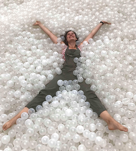 A woman smiles while lying in a bed of hundreds of white plastic balls. Her arms and legs are diagonal, making an X.