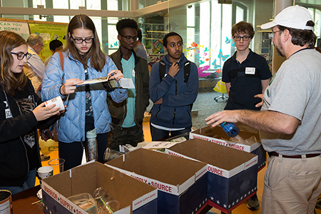 Five teens stand by a table with boxes on it holding recyclables. The museum’s Eric Hollinger is talking to the students.