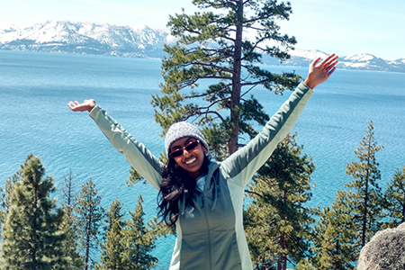 A woman wearing sunglasses and a ski cap standing in front of tree with water and mountains in the background.