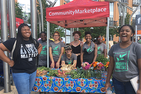 Eight teenagers stands near a table with fresh produce and flowers at an outdoor booth under a red canopy.