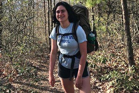A woman standing on a path in the woods, wearing a backpack.