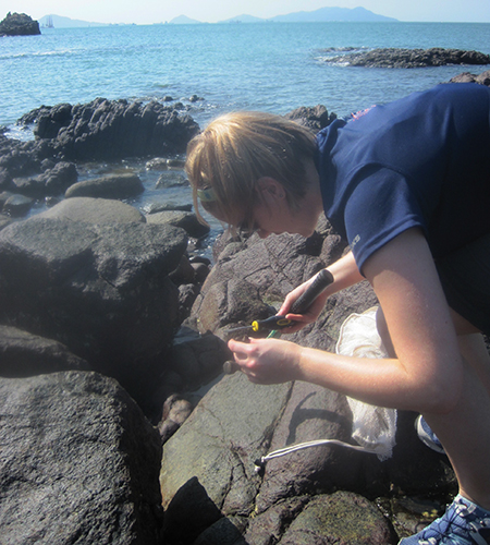 A woman holding a tool, examining something in her hand while bending down over some rocks near the water’s edge.