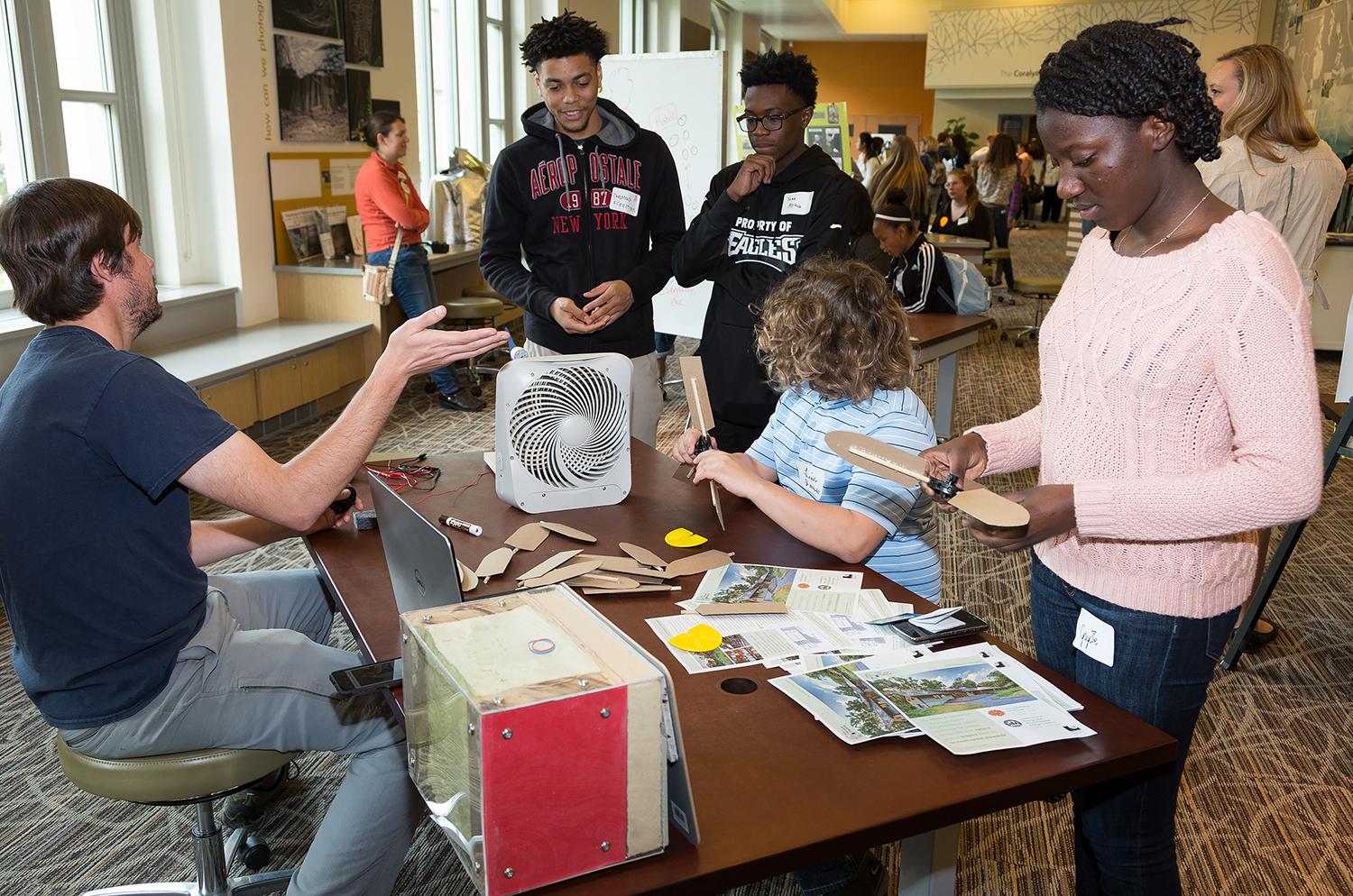 Three teens standing near a table while one sits at it. An adult is talking to them. Two teens are assembling something.