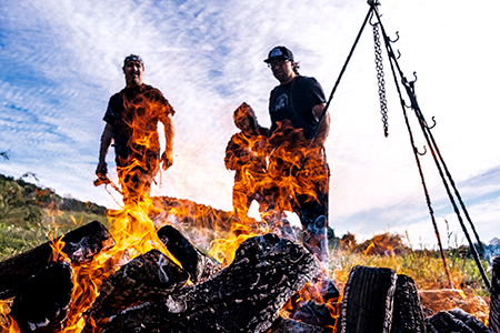 Three people standing near a stack of burning logs in a field. Near the fire is a triangular stand possibly used for cooking.