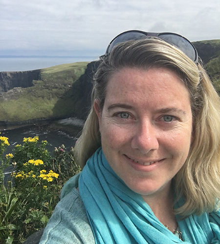 A woman with sunglasses on top of her head standing in front of a coastal scene with yellow flowers and green cliffs.