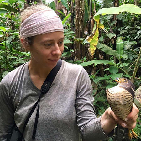 A woman standing in the woods or jungle, holding a bird in her left hand.