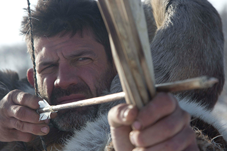 A bearded man holding a bow and arrow and pulling back the bow string while taking aim.