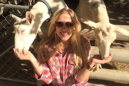 A woman with sunglasses holding out her hands to feed two goats, which are behind her in a pen.