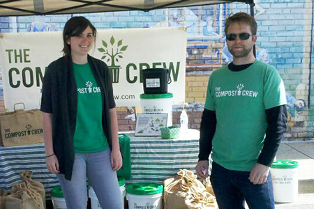 A man and woman standing under a canopy, wearing green “The Compost Crew” t-shirts. Behind them is a table display.