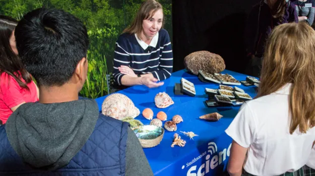 Zoologist Ellen Strong gestures with one hand as she talks to young students gathered around a table of museum specimens of snails and other mollusks.