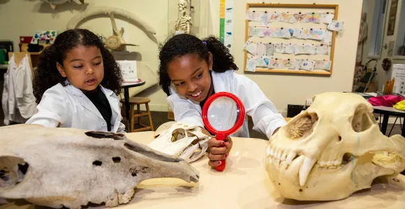 Two young girls in white lab coats use a hand lens to examine models of fossils at a table in Q?rius jr. – a discovery room.