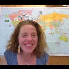 Jen Kretser, a light-skinned woman with shoulder-length brown hair, sitting in front of a world map on the wall behind her