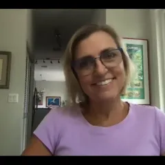 Video-conference image of a smiling woman with short blonde hair, glasses and a lavender shirt