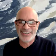 A bald man with glasses and gray beard, wearing a navy blue top. Behind him is a background of a blue and white painting.