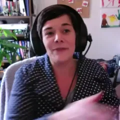 A woman with dark hair and headphones, sitting in an office with a plant and bookshelf behind her.