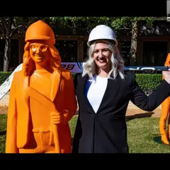 A woman with blonde hair, wearing a black blazer and a white hard hat stands next to an orange statue of herself