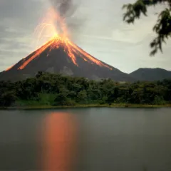 Volcano erupting with lava streaming down the side. A forest is at its foot and the eruption is reflected in a dark lake. 