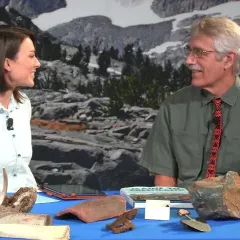 Maggy Benson and Dr. Bill Fitzhugh are seated at a table holding some rocks, antlers, tools, and a book.