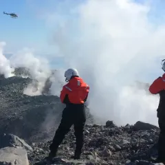 Three people wearing white helmets and red jackets stand atop a steaming ridge as a helicopter hovers nearby.