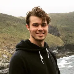 photo of young man with brown hair smiling in front of green hills and water