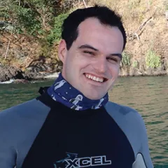 A man in a wetsuit smiling in front a body of water