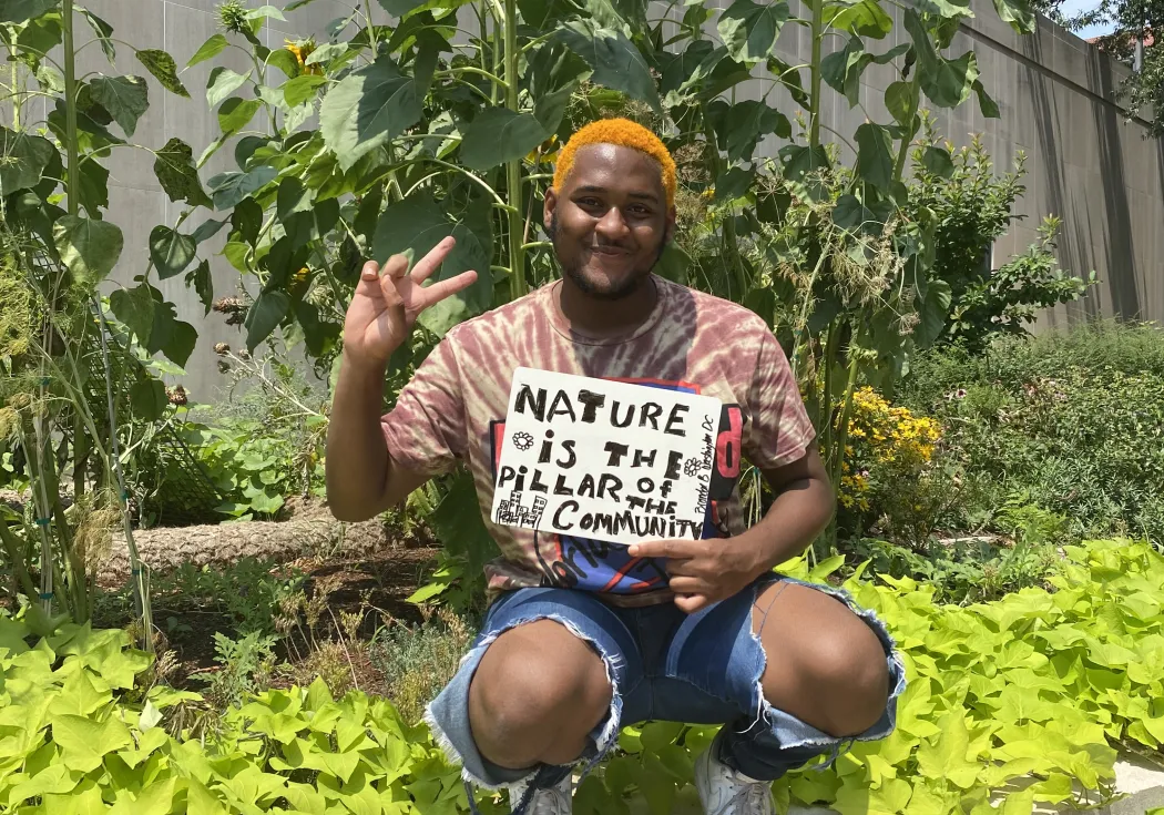 a black man with orange hair kneeling under giant sunflower with a sign reads nature is the pillar of community