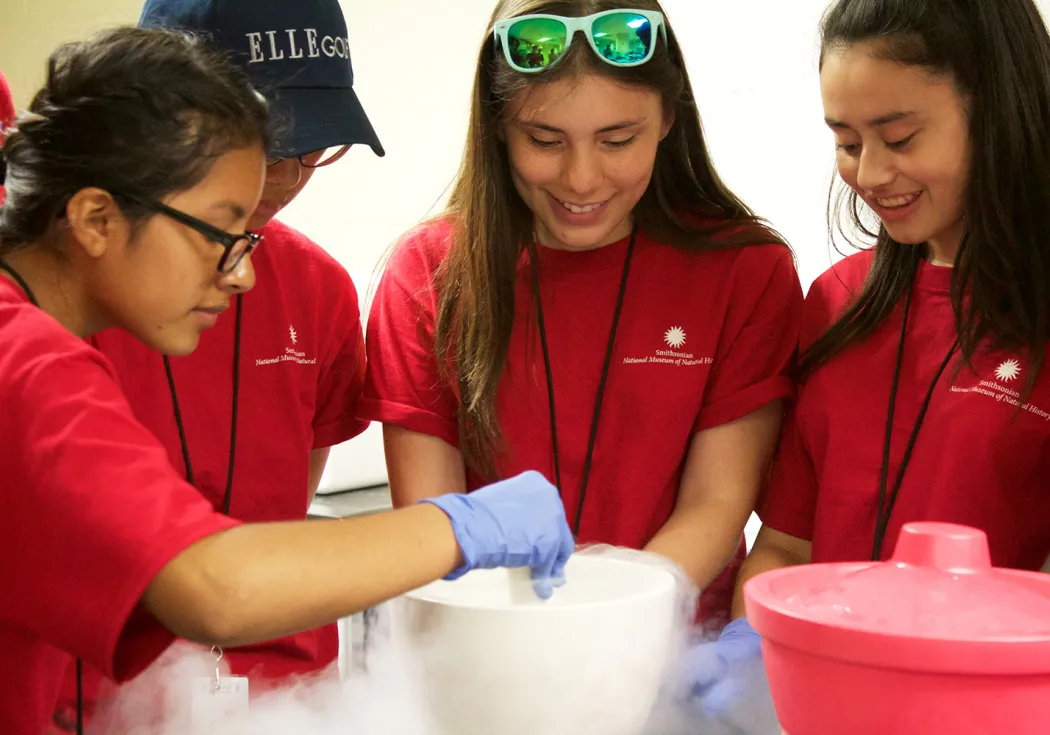 Four teenagers in red t-shirts and blue gloves stand around a white bucket with smoke or steam coming out of it.