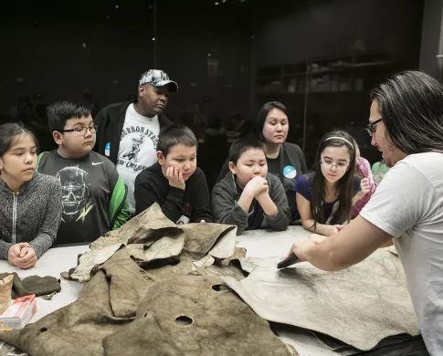 School students around a table, watching a man softening a moose hide with a stone scraper
