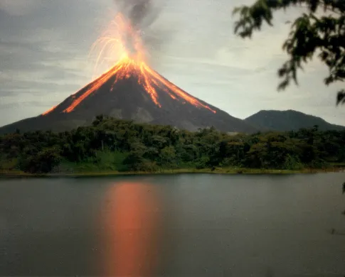 Volcano erupting with lava streaming down the side. A forest is at its foot and the eruption is reflected in a dark lake. 