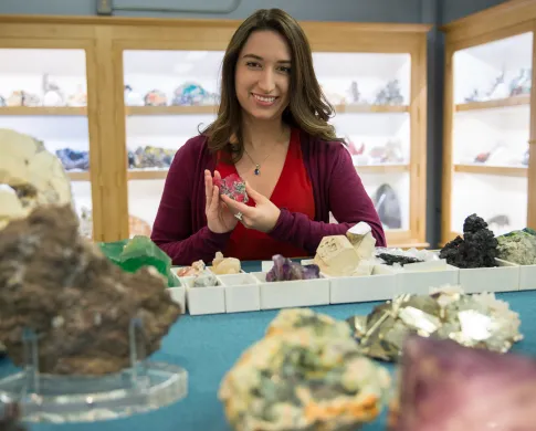 A woman holding a reddish-pink mineral while standing at a table that has many other minerals, and gems on it.