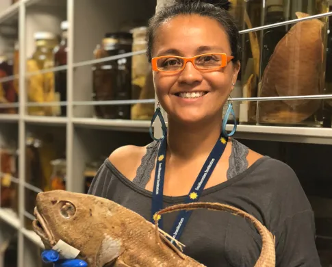 A woman holding a fish specimen in front of jars of other fish specimens.