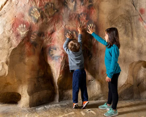 Two children put their hands over handprints on the wall of a model cave in the Human Origins exhibit.