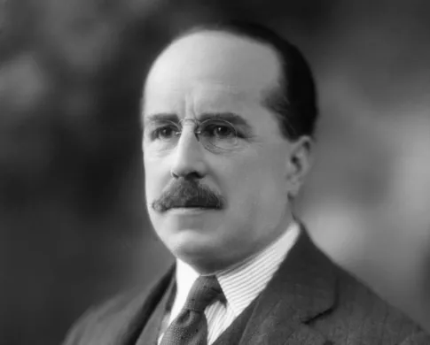 A serious, light-skinned man with glasses and a mustache looks past the camera