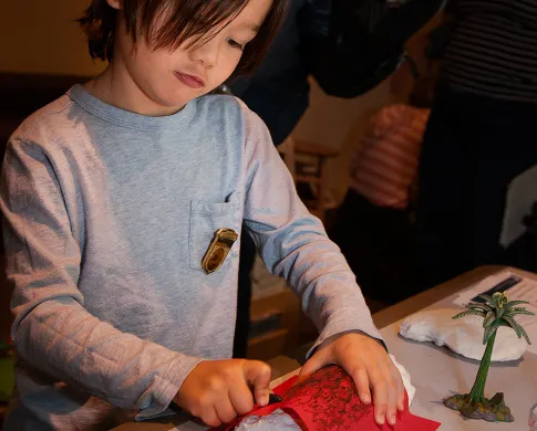 A child holding a red piece of paper on top of a rock or fossil while rubbing a dark crayon on the paper