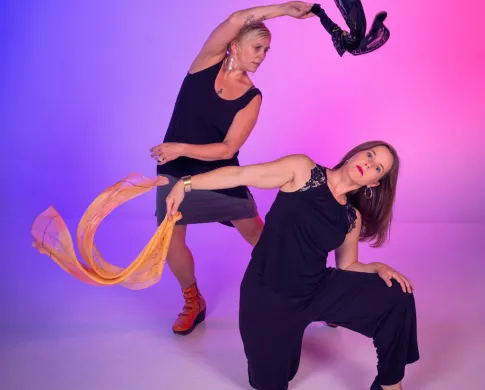 Two women dancing and waving scarves against a purple-pink background.