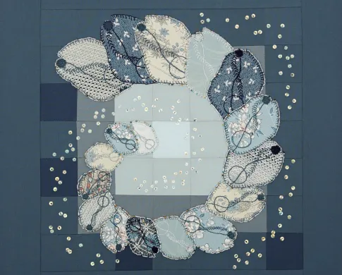Quilt with squares of different shades of blue and gray, and embroidery of a spiral chain of rectangles of increasing size. The rectangles each have curved corners, a large black dot inside, and are beige, gray, and different shades of blue.