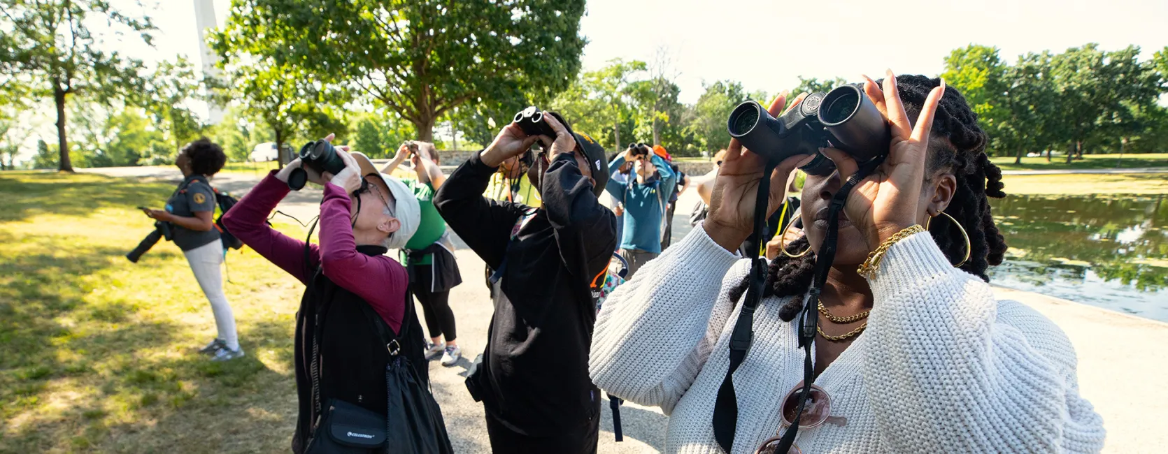 A group of about eight people, including some medium- and light-skinned people, standing on a path on the National Mall and looking up through binoculars. One woman is holding a camera. The Washington Monument is in the background along with some green, leafy trees.