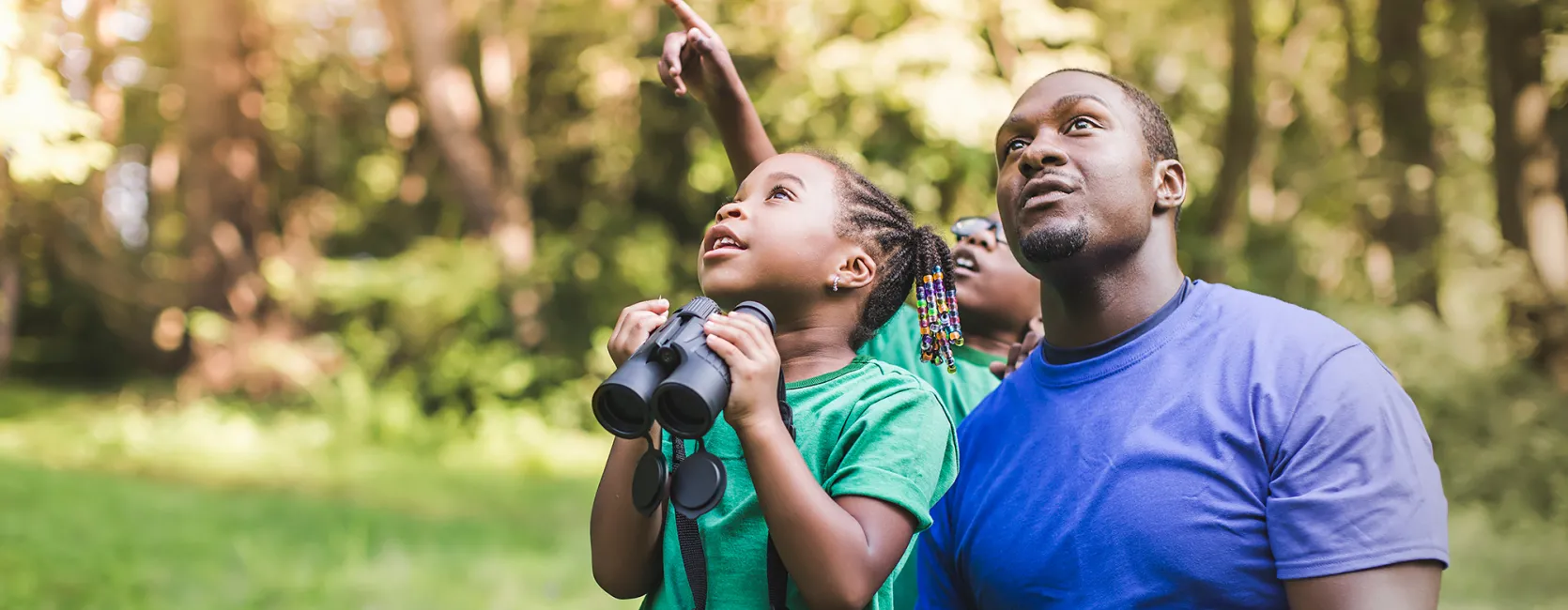 A dark-skinned man and his young daughter and son in front of some trees, looking up as if birdwatching. The girl holds binoculars and the boy is pointing diagonally up.