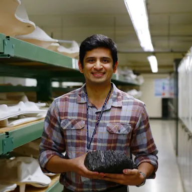 Advait Jukar in front of shelves holding jacketed fossils and holding a fossil tooth