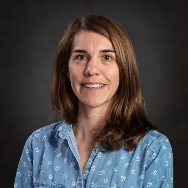 A woman with brown hair in a blue and white patterned button up shirt smiling for a professional headshot photograph 