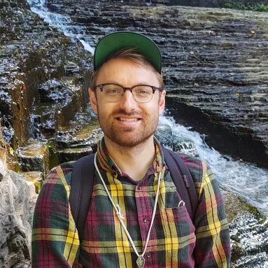 Male with beard wearing hat, glasses, neckless, and a multicolor plaid shirt