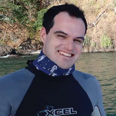 A man in a wetsuit smiling in front a body of water