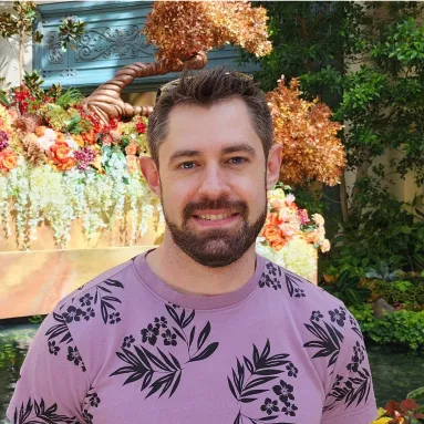 A bearded, light-skinned man in a floral lavender shirt in front of a flowerbed