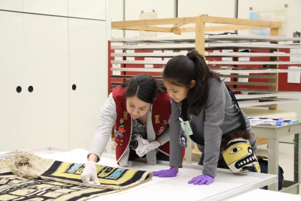 Two people examining a Chilkat blanket with yellow and blue colors.